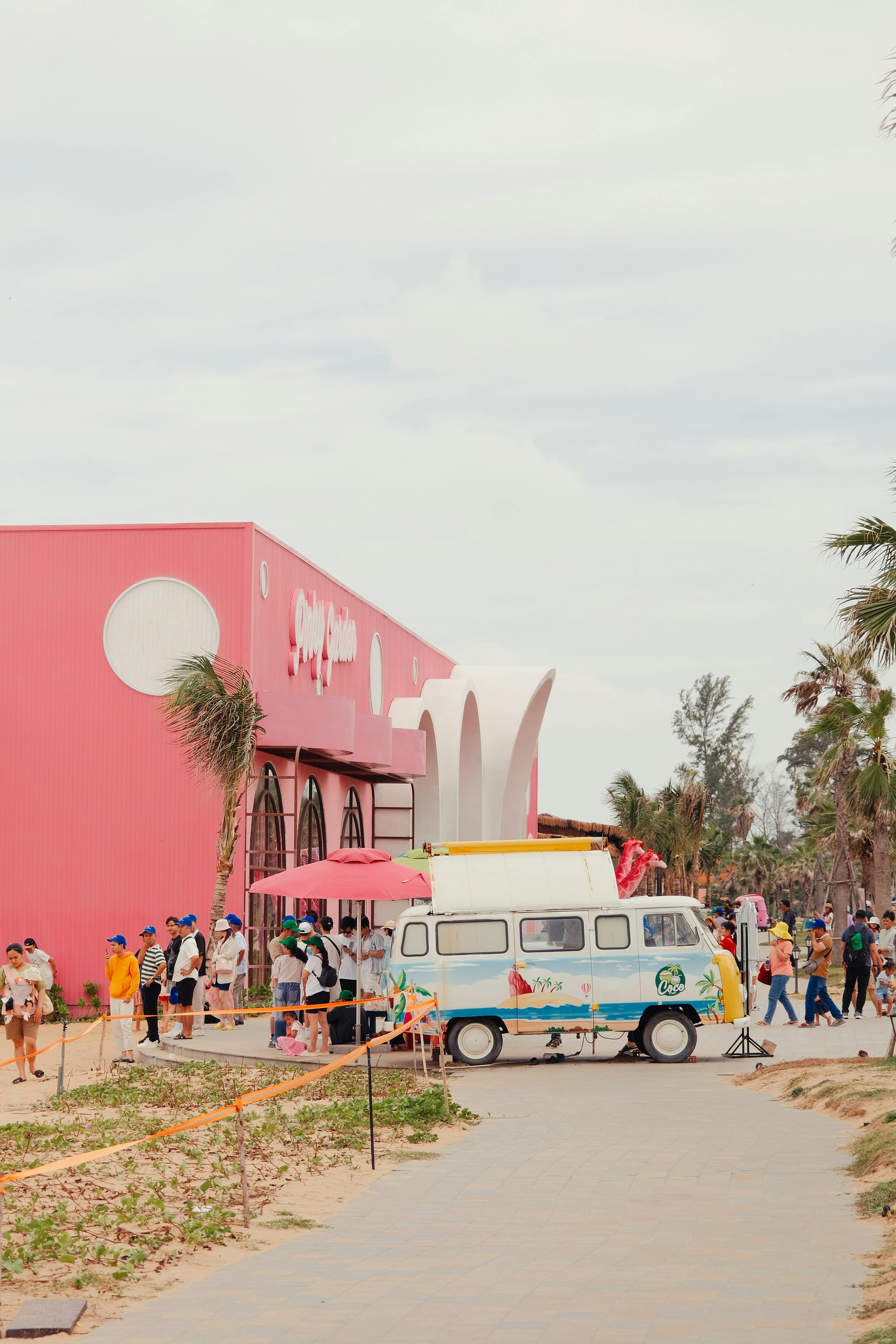 A colorful scene with a retro van parked by a vibrant pink building, surrounded by people enjoying the outdoors.