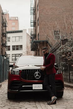 Stylish man in formal attire leaning on a luxury car in a city alleyway.