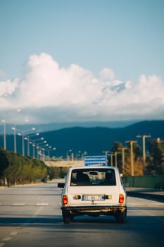 Classic white car driving on an empty road against a backdrop of mountains and blue sky.