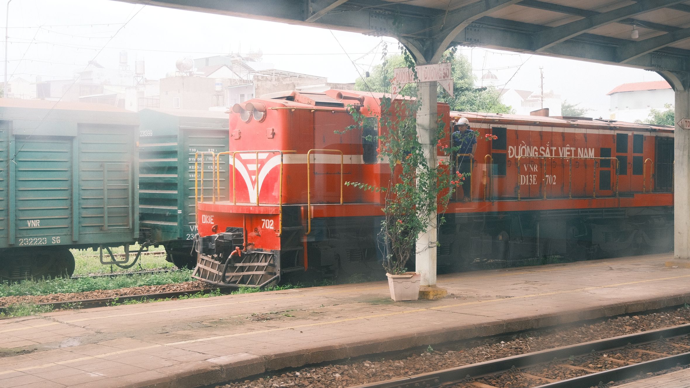 A red train parked at a Vietnamese urban station under a covered platform. Industrial and urban feel.