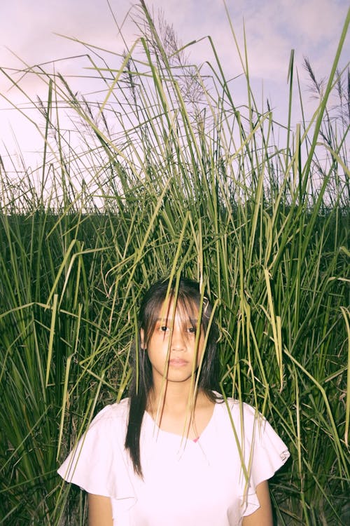 Emotionless female teen in casual wear with dark hair looking at camera standing near thin pointed growing grass in countryside field under sky in summer evening