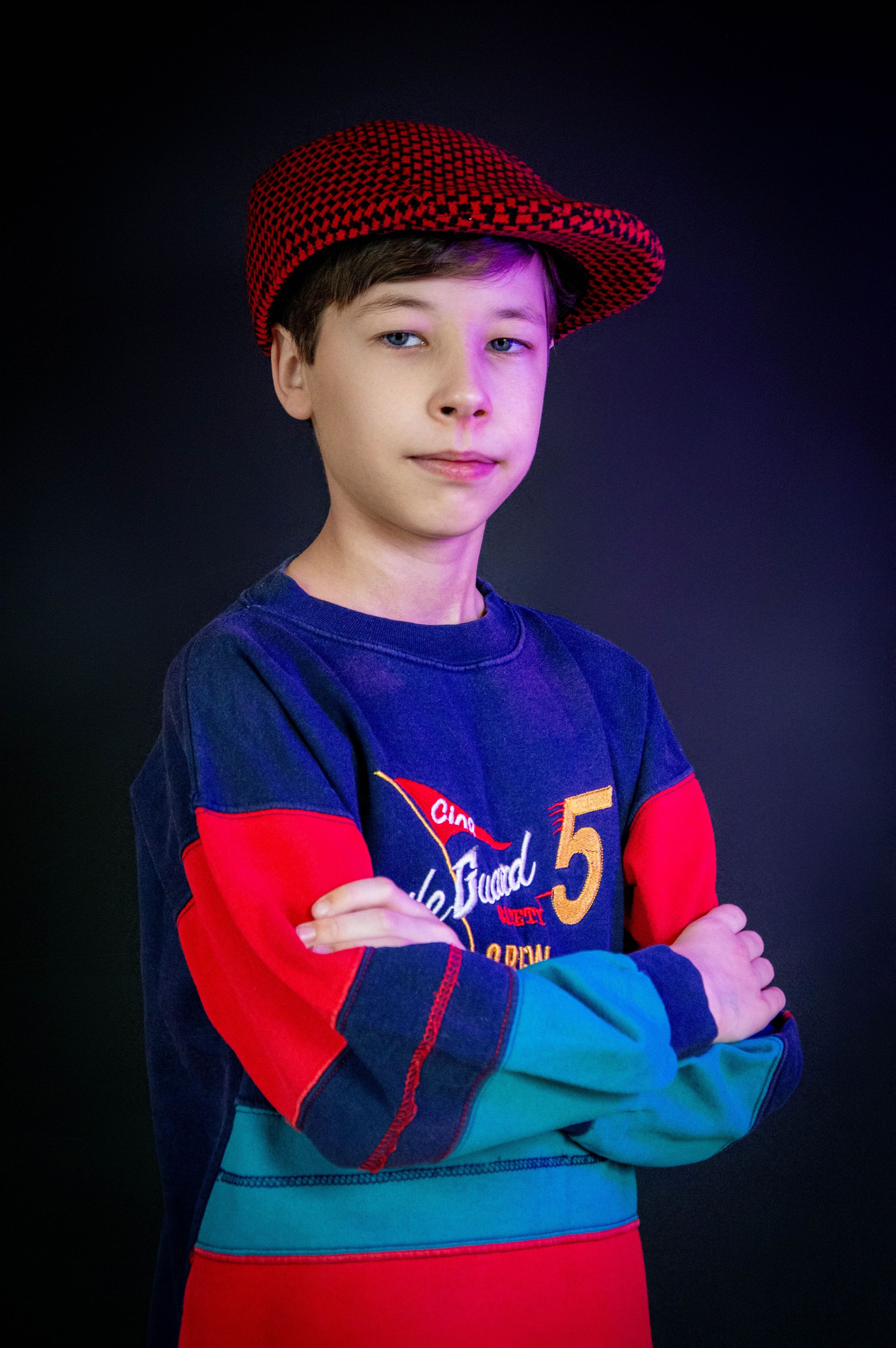 Young teenager in vibrant clothes and red cap crossing arms against dark background.