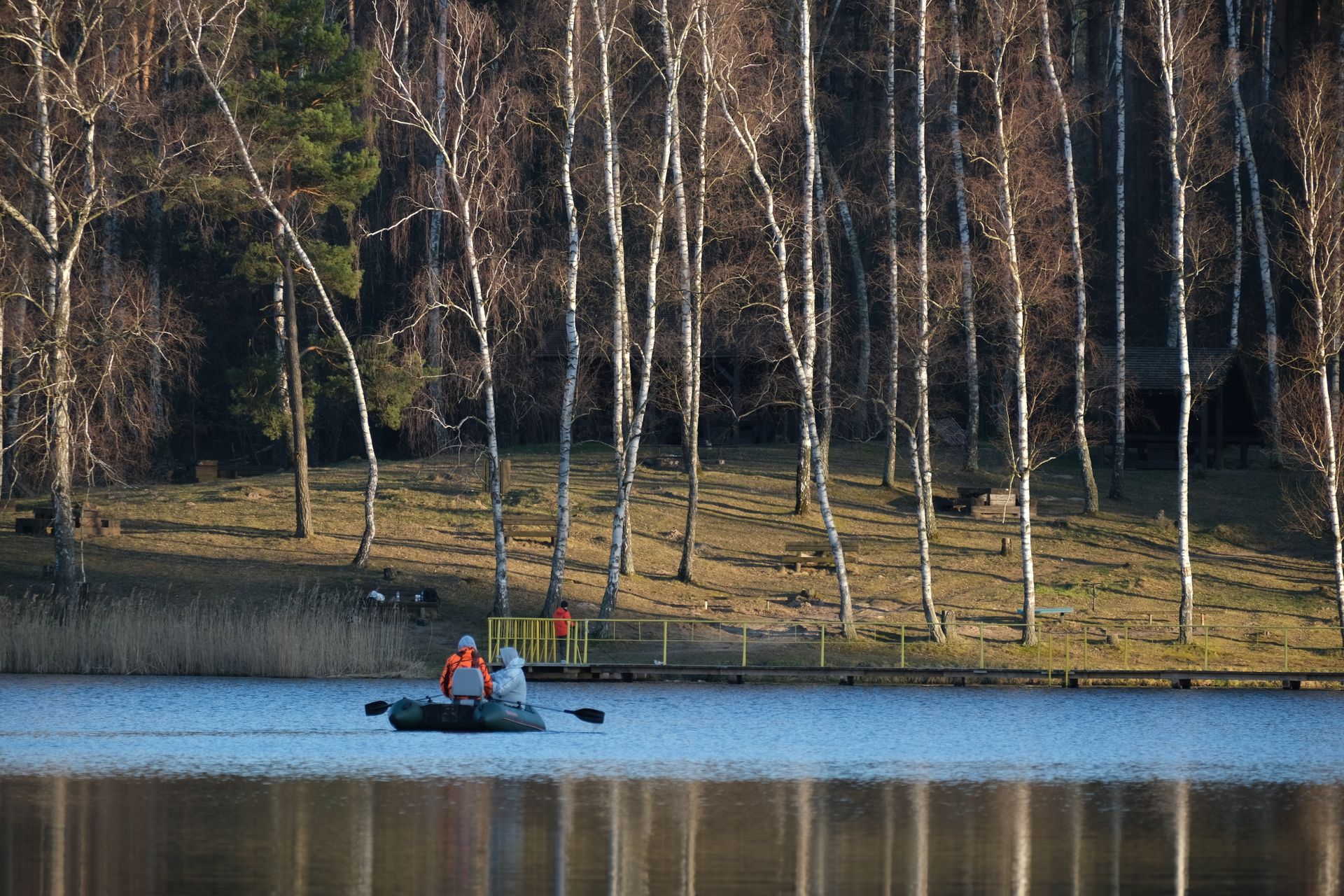 Two people rafting on a calm river surrounded by leafless birch trees in a tranquil setting.