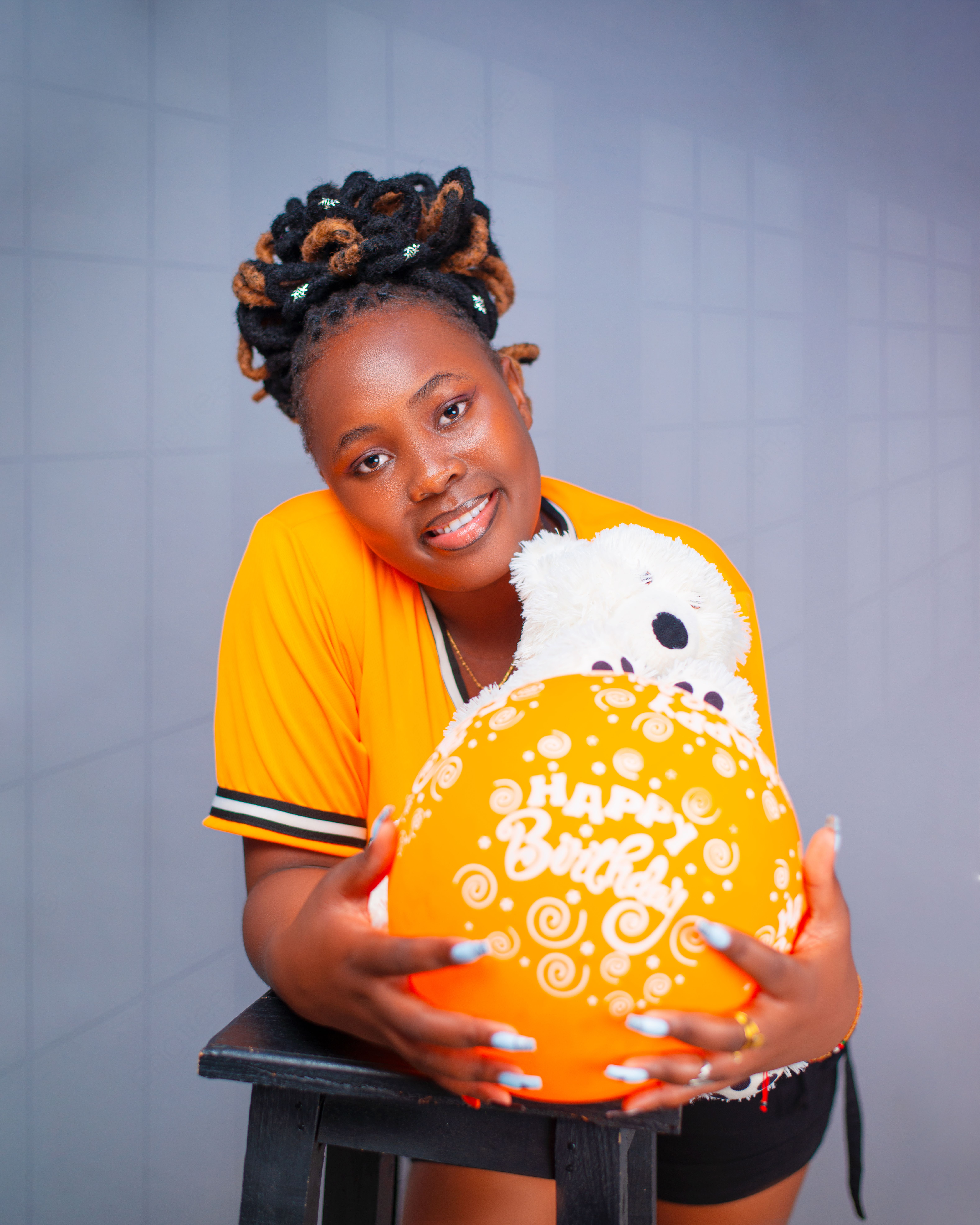 A smiling woman holding a vibrant birthday balloon and a teddy bear indoors.