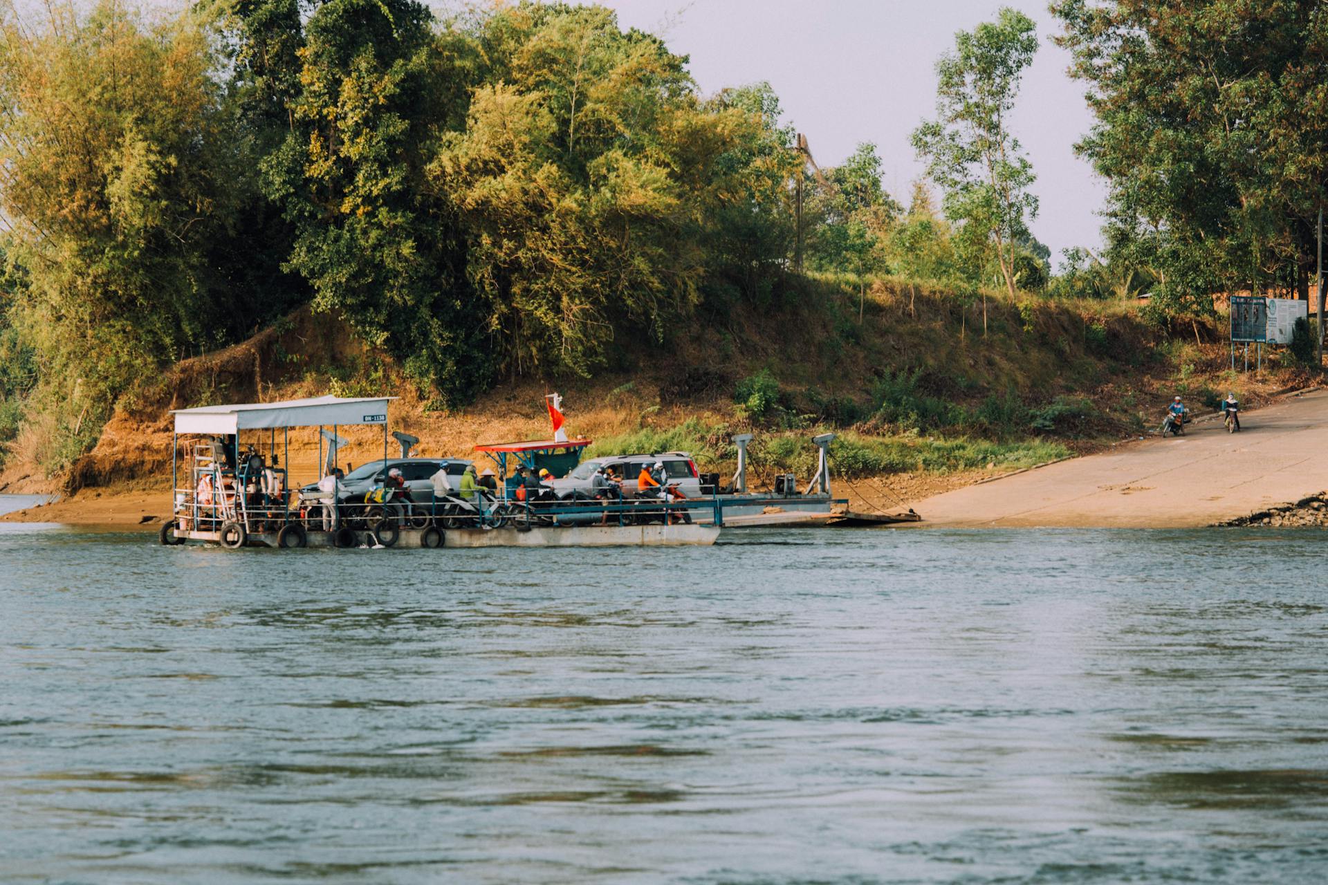 A vibrant river ferry transporting vehicles and passengers, set against a lush green backdrop.