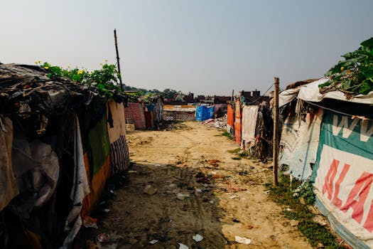 一条尘土飞扬的乡村小路两侧，搭着覆盖防水布和标志的临时避难所