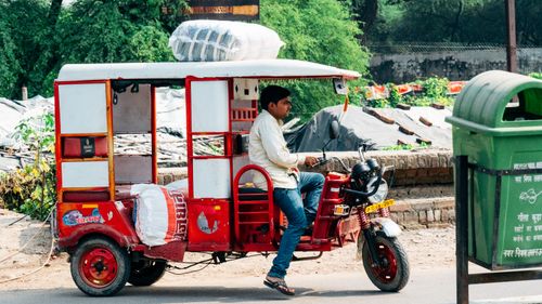 Free A man driving a red bicycle rickshaw loaded with goods through an urban street. Stock Photo