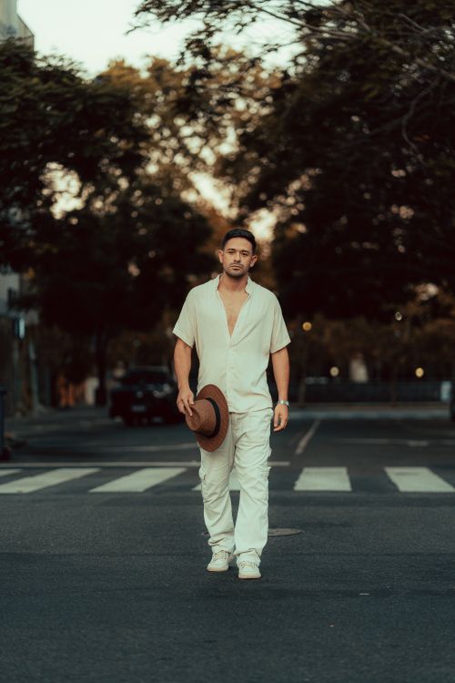 Free A fashionable young man in white attire walks confidently on a city street during dusk, holding a hat. Stock Photo