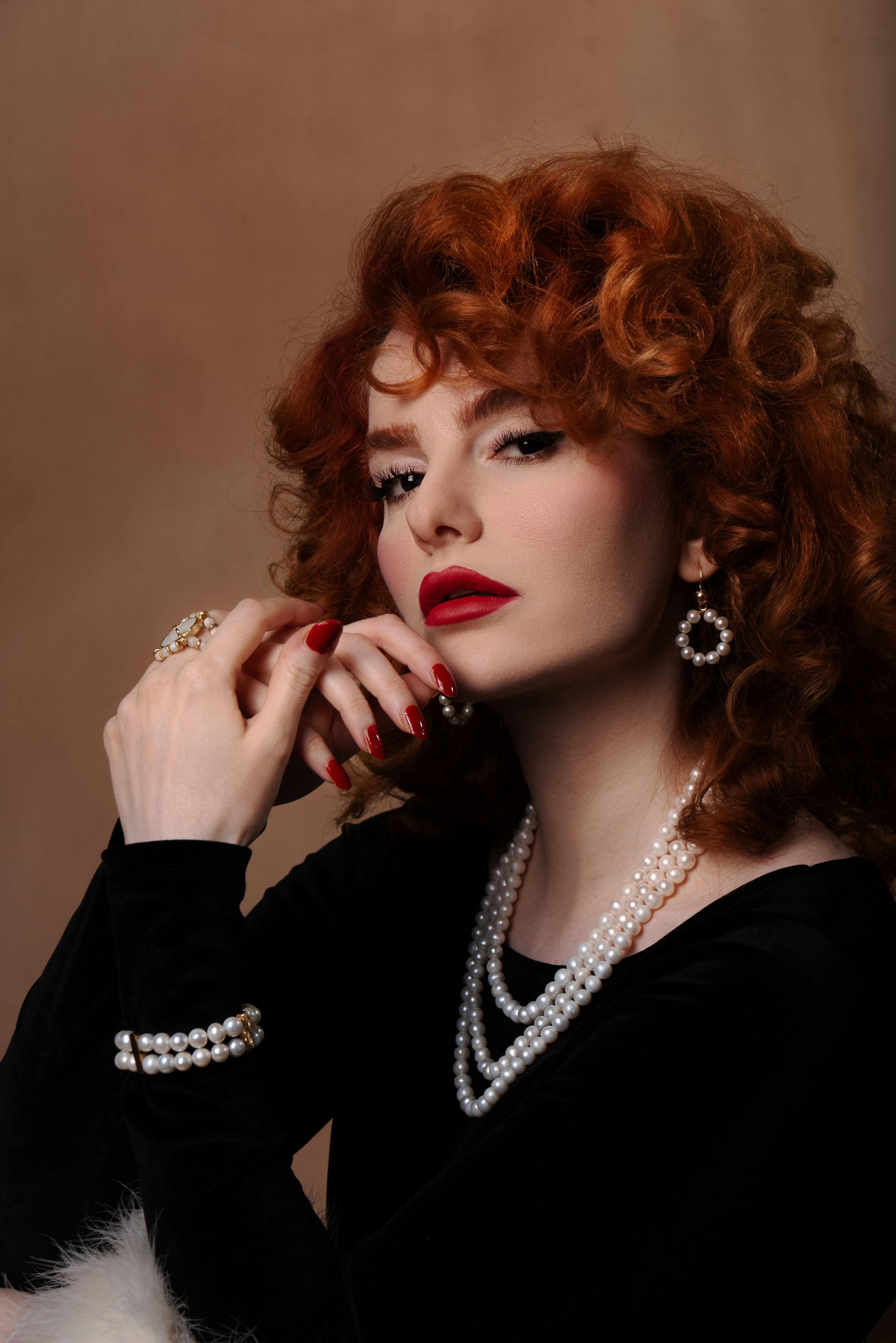 Sophisticated woman with red hair and pearl jewelry against a brown backdrop.
