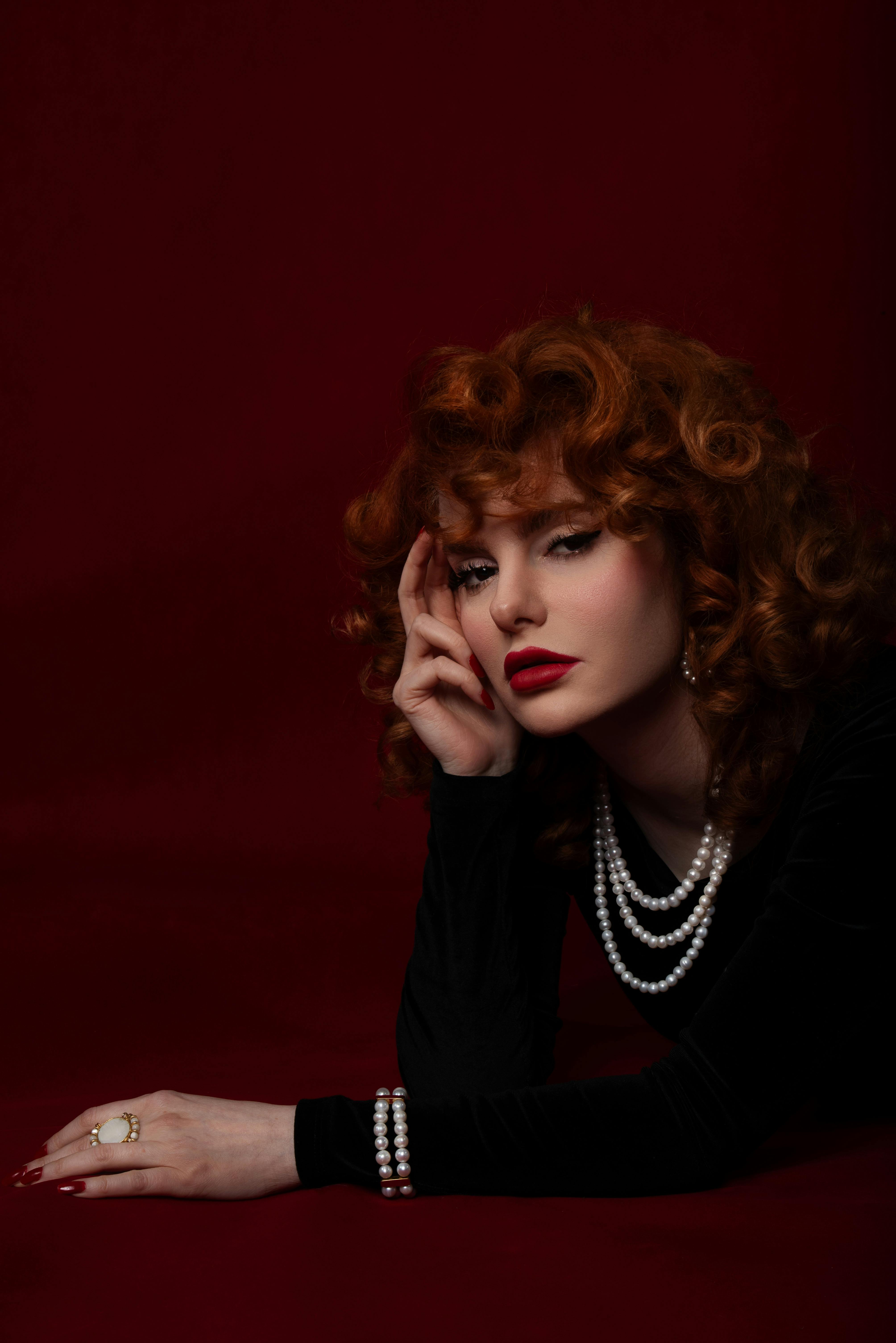 Stylish woman with red curls and pearls, posing on rich red background.