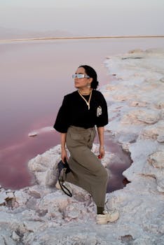 Stylish woman in sunglasses poses by a pink salt lake, showcasing modern fashion.