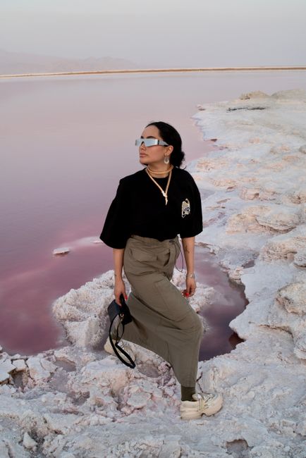 Stylish woman in sunglasses poses by a pink salt lake, showcasing modern fashion.