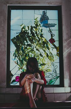 一名女子坐在色彩斑斓的玻璃窗旁沉思，窗上投下缤纷的倒影