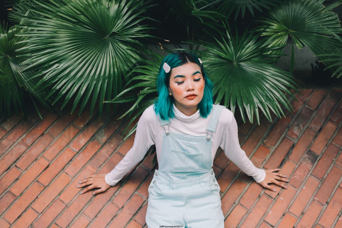 Free Fashionable woman with teal hair sitting against a backdrop of lush green leaves. Stock Photo