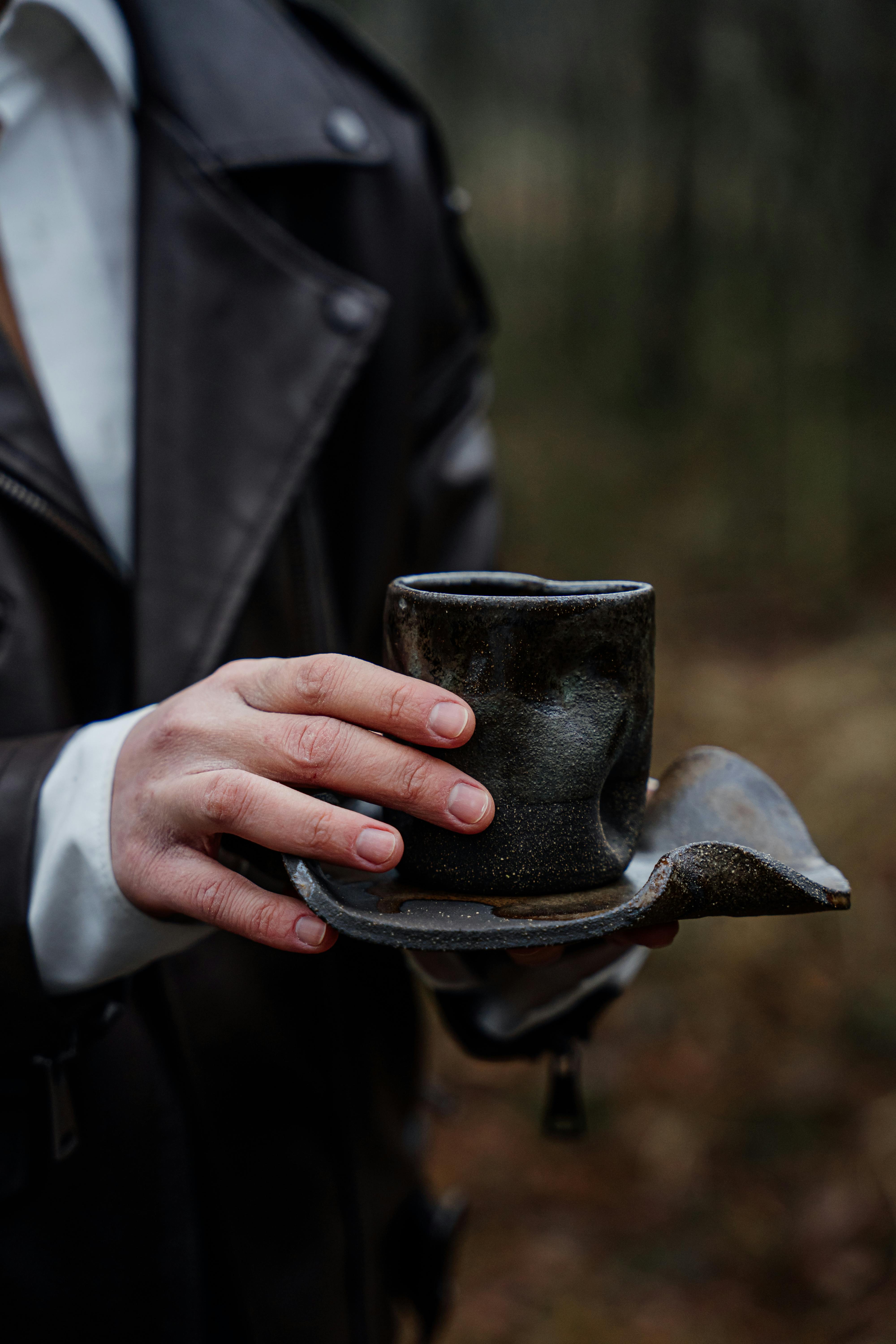 Close-up of a person holding a rustic handmade ceramic mug with saucer outdoors.