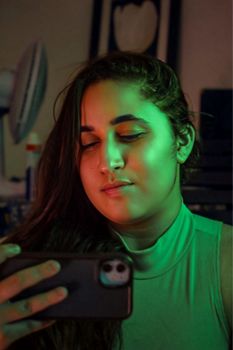 A young woman uses her smartphone in a dimly lit room with green ambient lighting.
