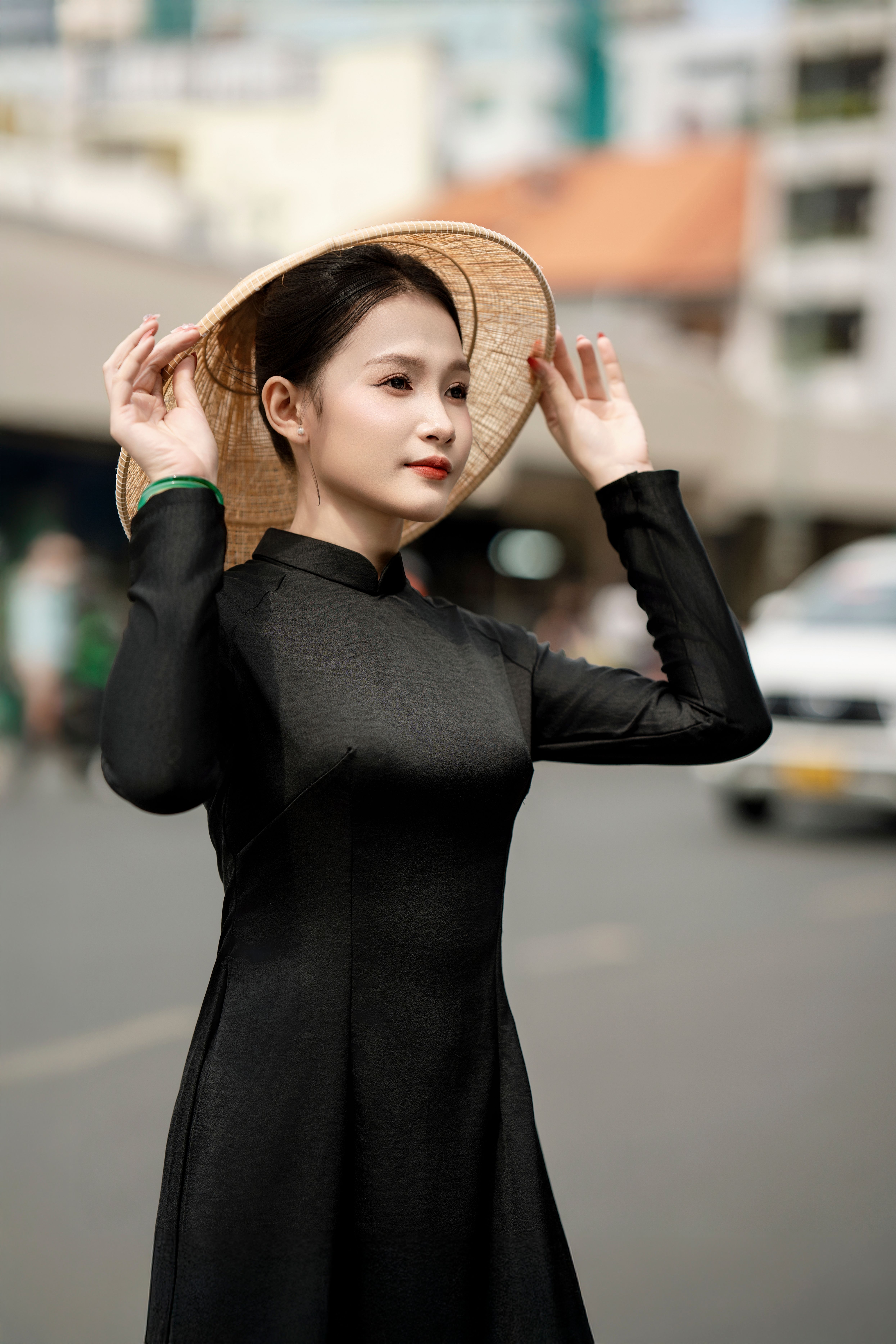 Elegant woman in a black Ao Dai with a hat in an urban setting, exuding cultural grace.