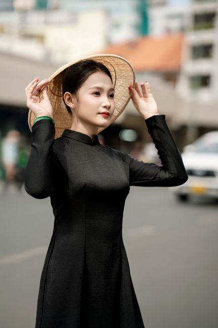 Elegant woman in a black Ao Dai with a hat in an urban setting, exuding cultural grace.