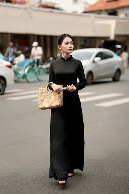 Elegant woman in black ao dai with straw bag walking down urban street.