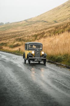 Classic vintage car driving on a wet, picturesque country road amidst golden fields.