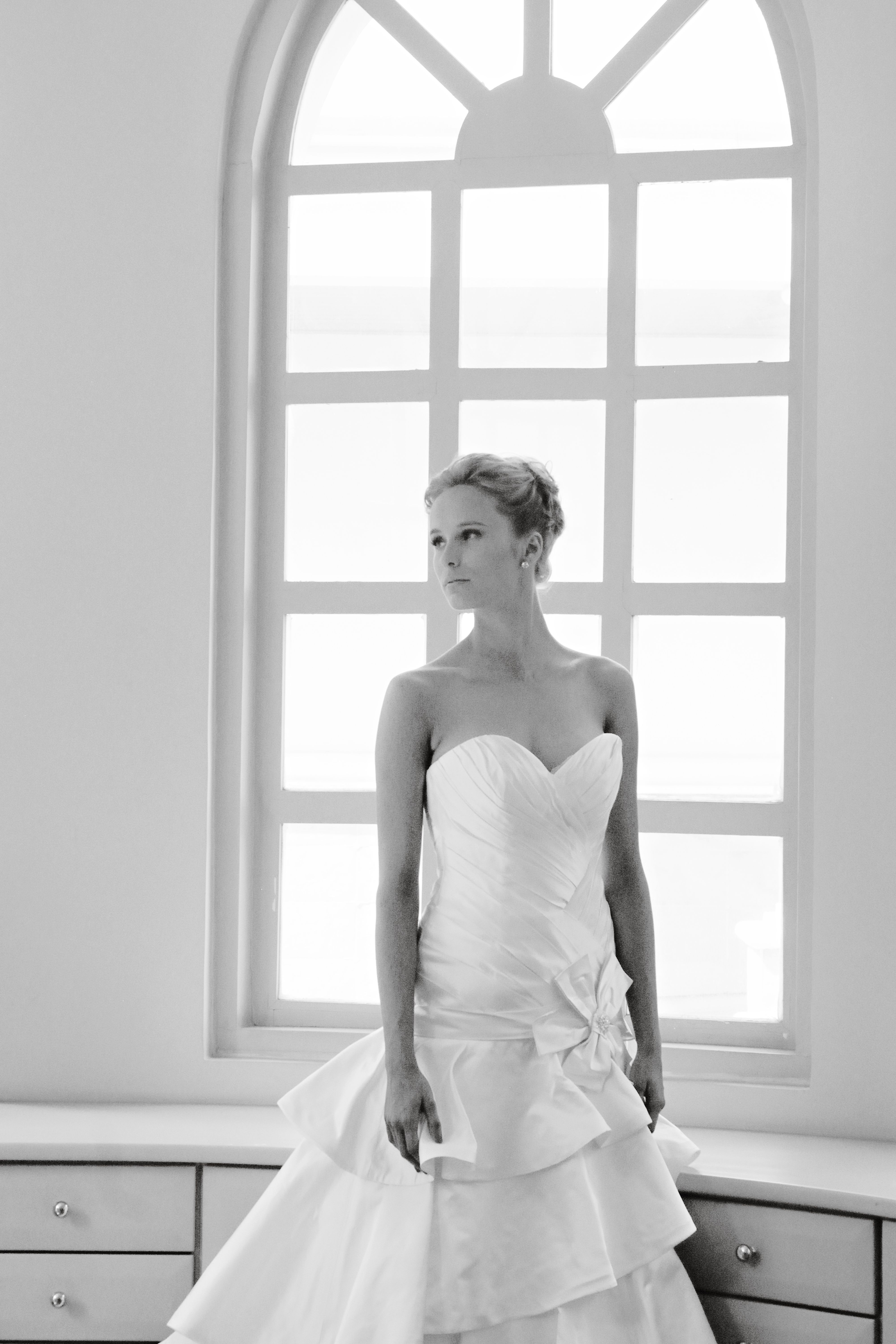 A striking black and white portrait of a bride standing by a window in a beautiful dress.