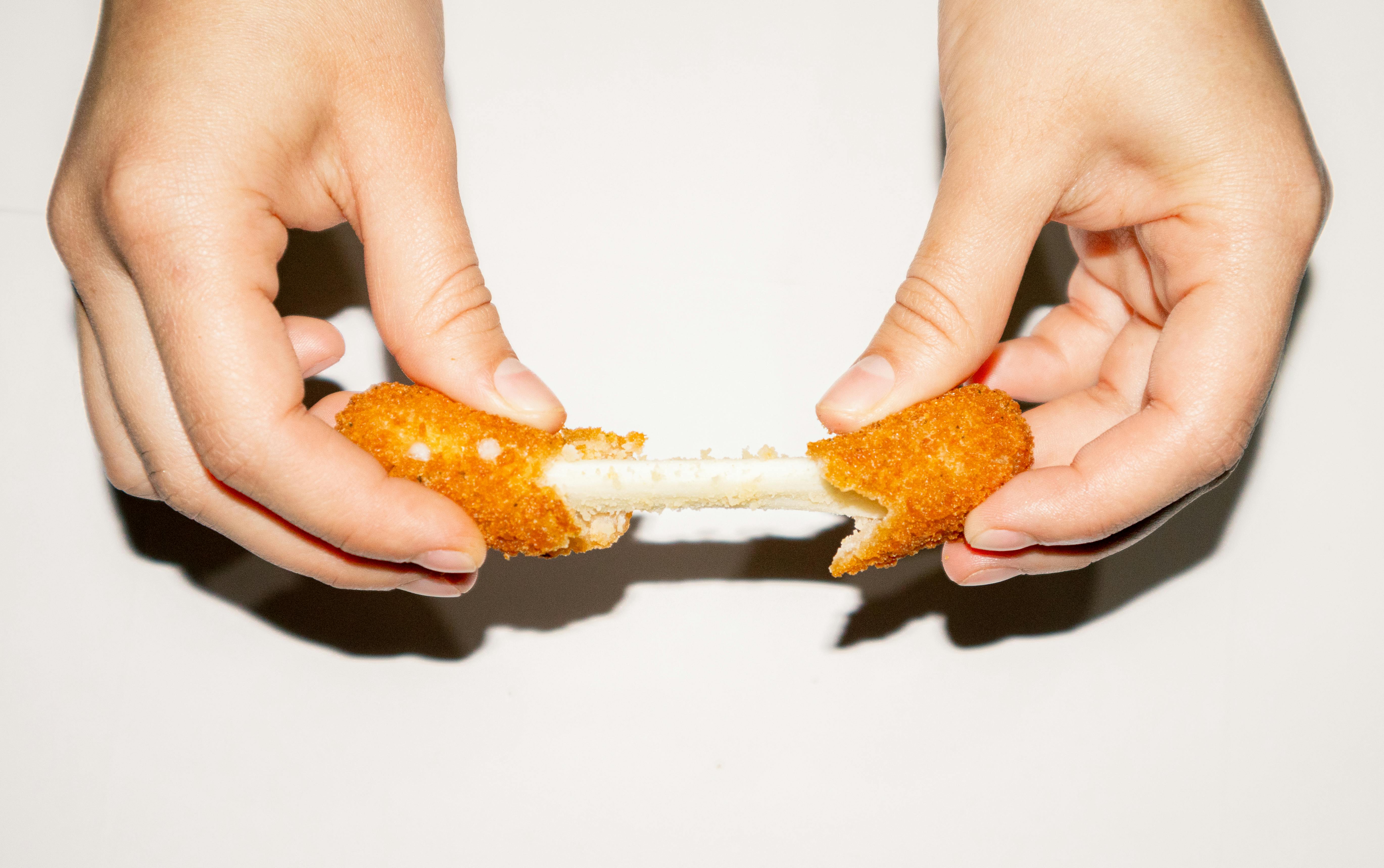 Hands stretching melted cheese from a crispy mozzarella stick on a white background.