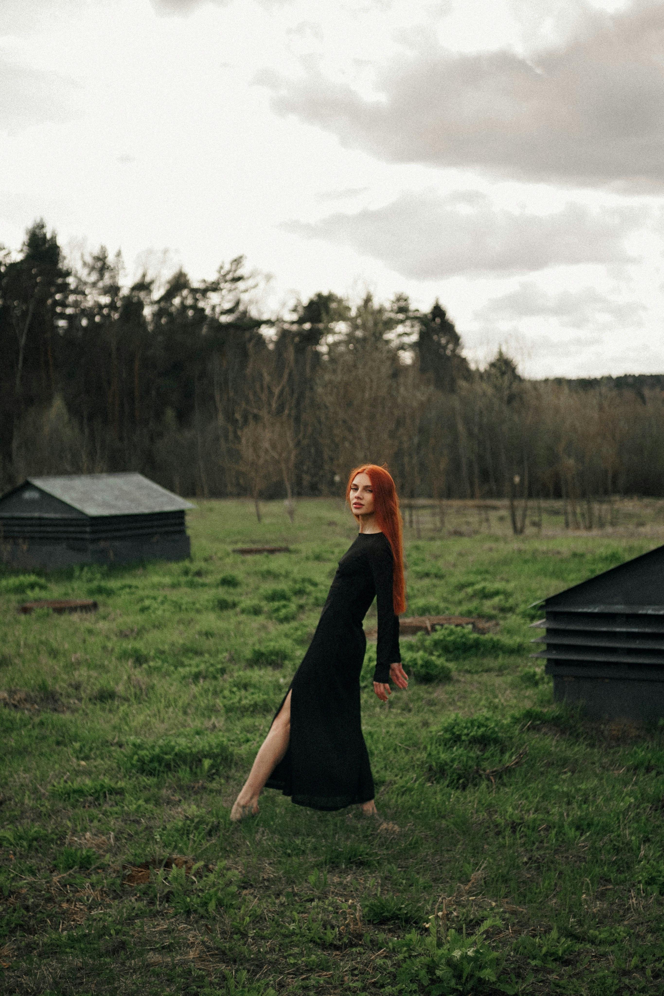 Kostenlos Eine Frau in einem schwarzen Kleid steht elegant auf einer Wiese mit Wald im Hintergrund. Stock-Foto