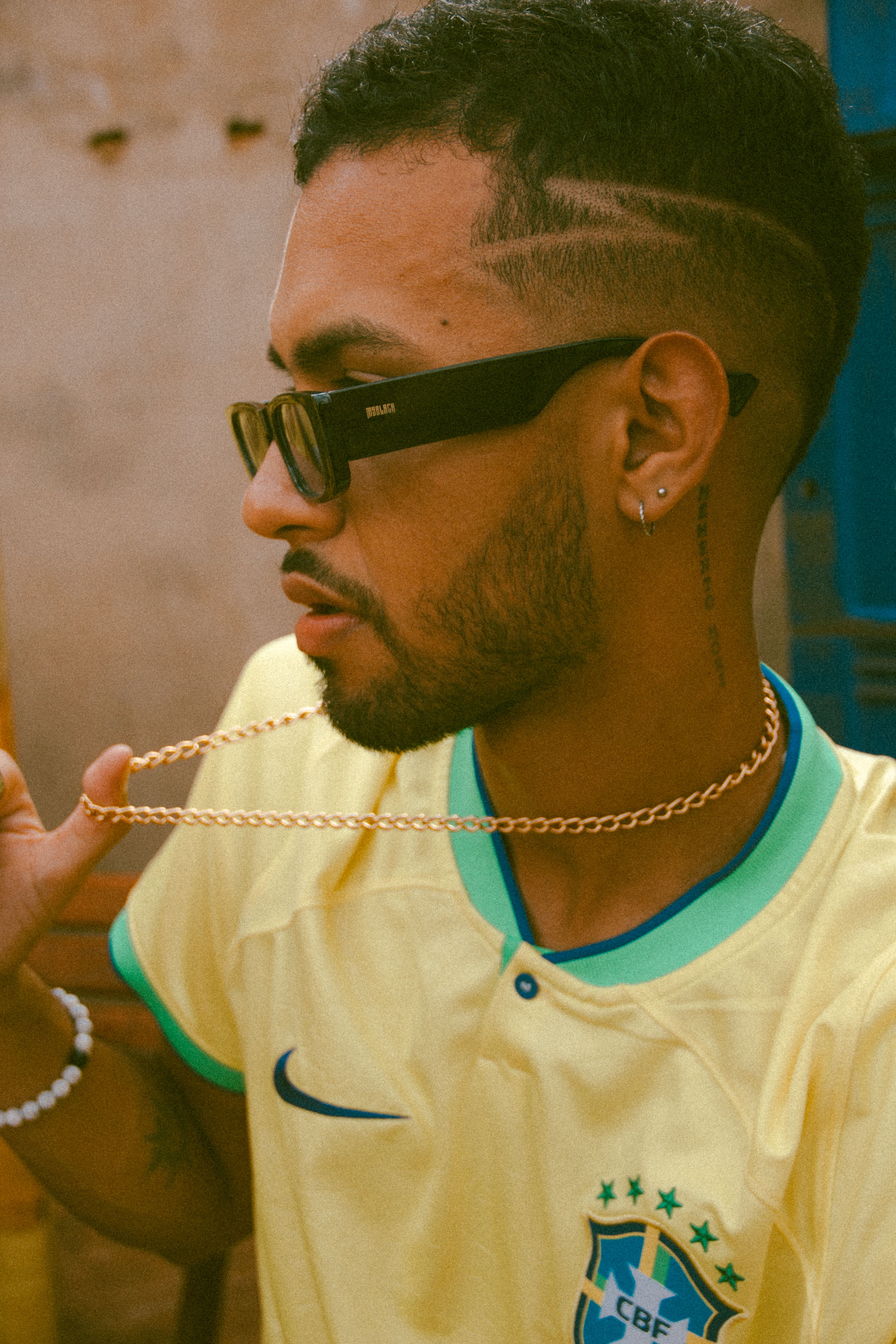 Young man wearing sunglasses and Brazil soccer jersey holding gold chain outside.