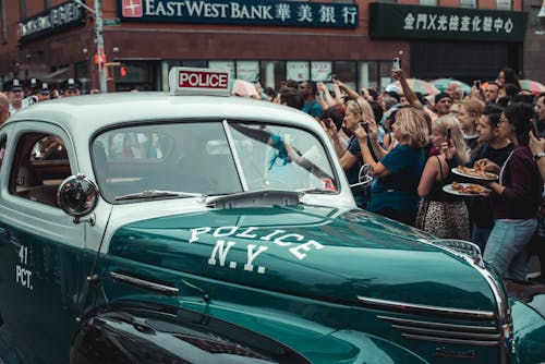 Gratis Una Folla Di Persone Raccolte Intorno A Un'auto Della Polizia Vintage Verde Nella Strada Della Città Foto a disposizione