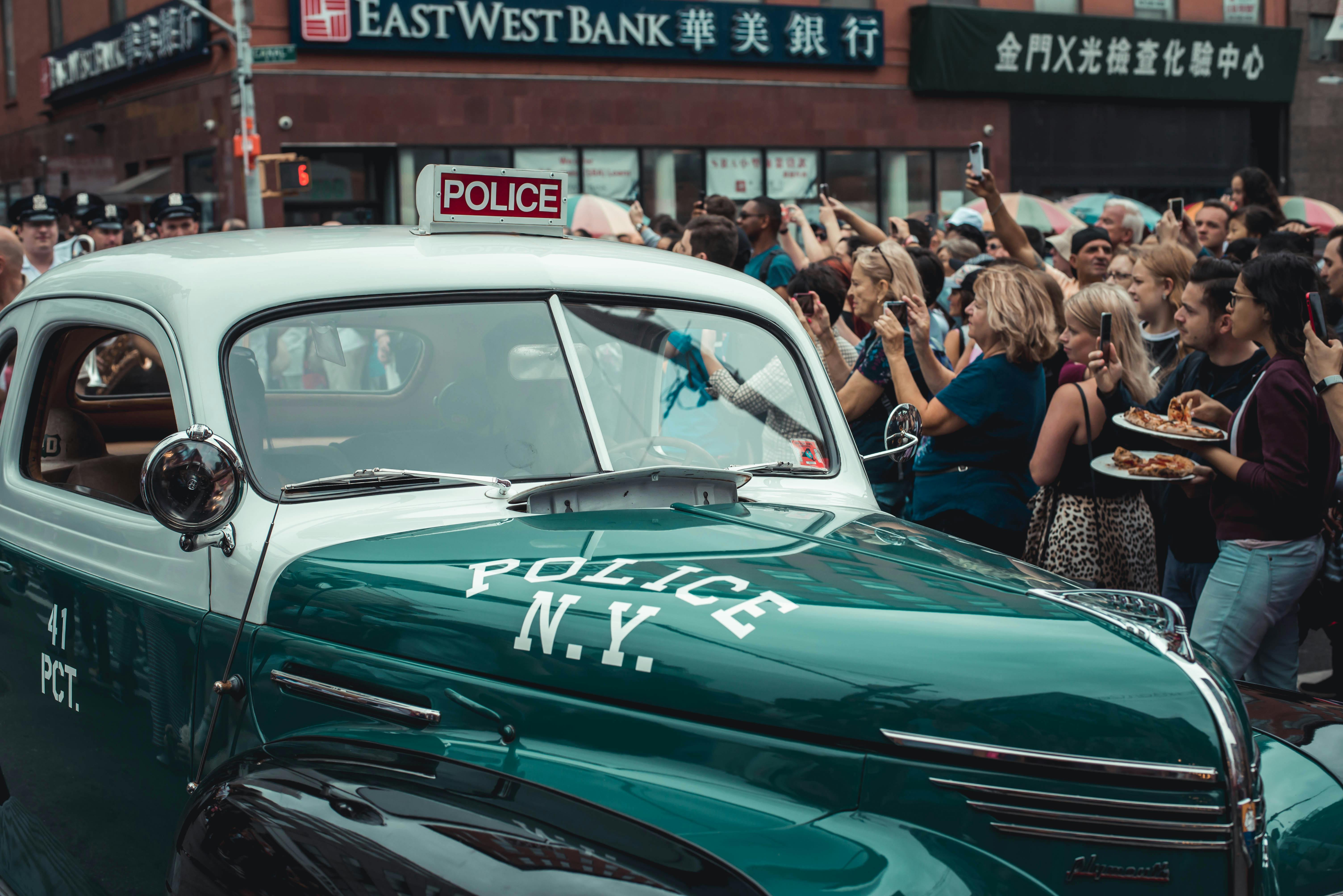 A Crowd Of People Gathered Around A Green Vintage Police Car In The