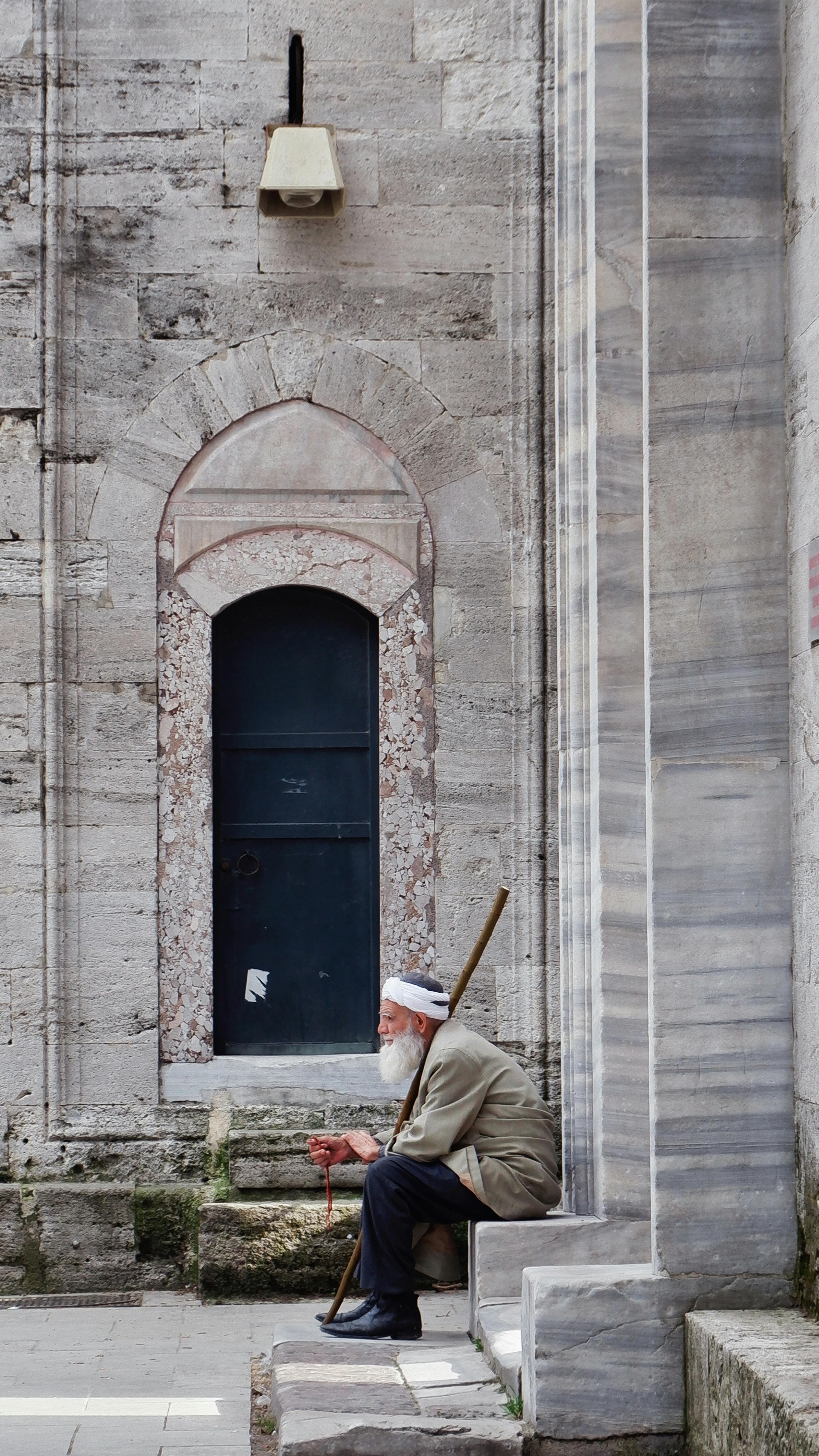 An elderly man sits by a historic stone wall, holding a staff, reflecting on life.