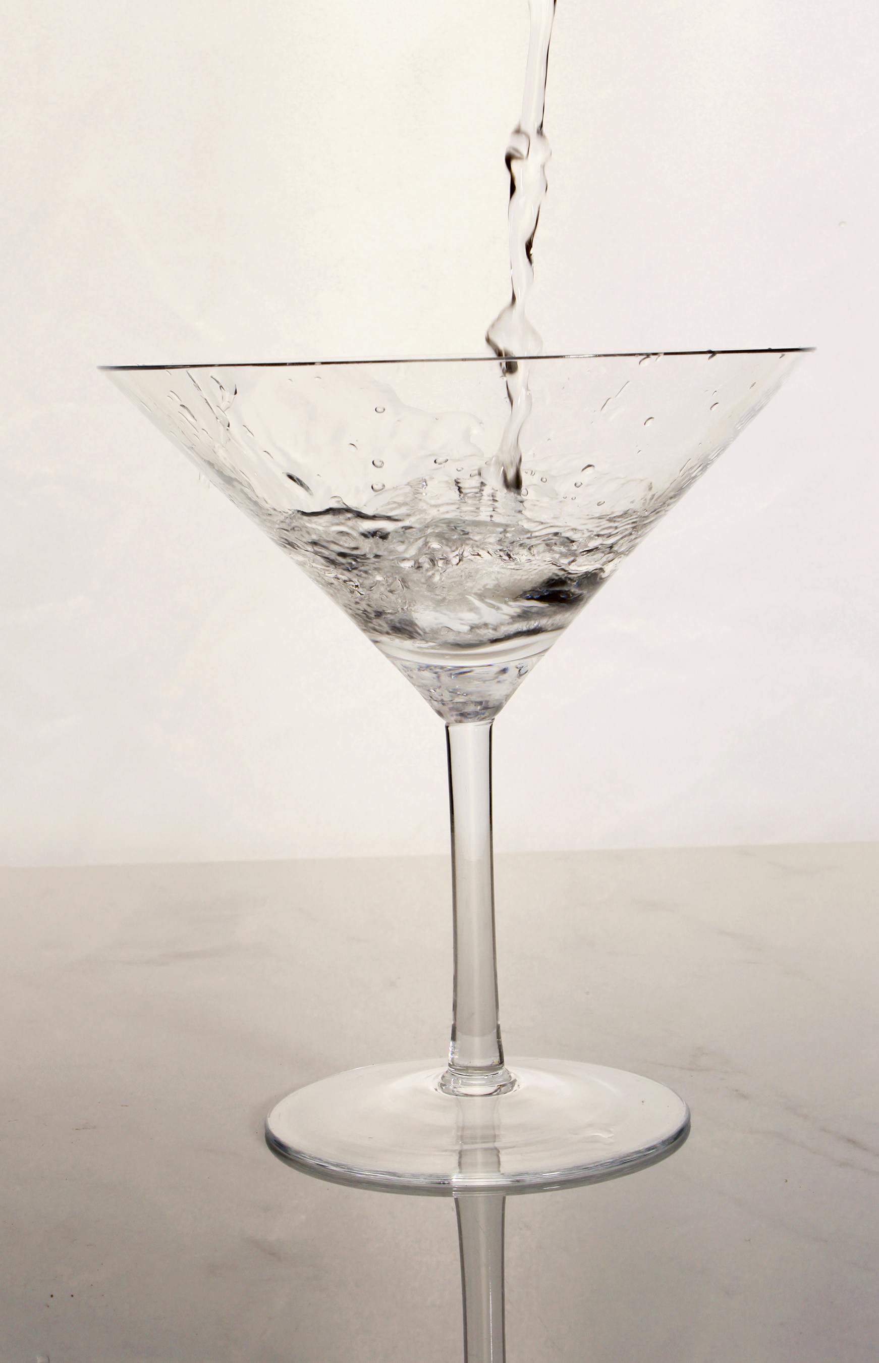 Close-up of water being poured into an elegant martini glass against a neutral background.