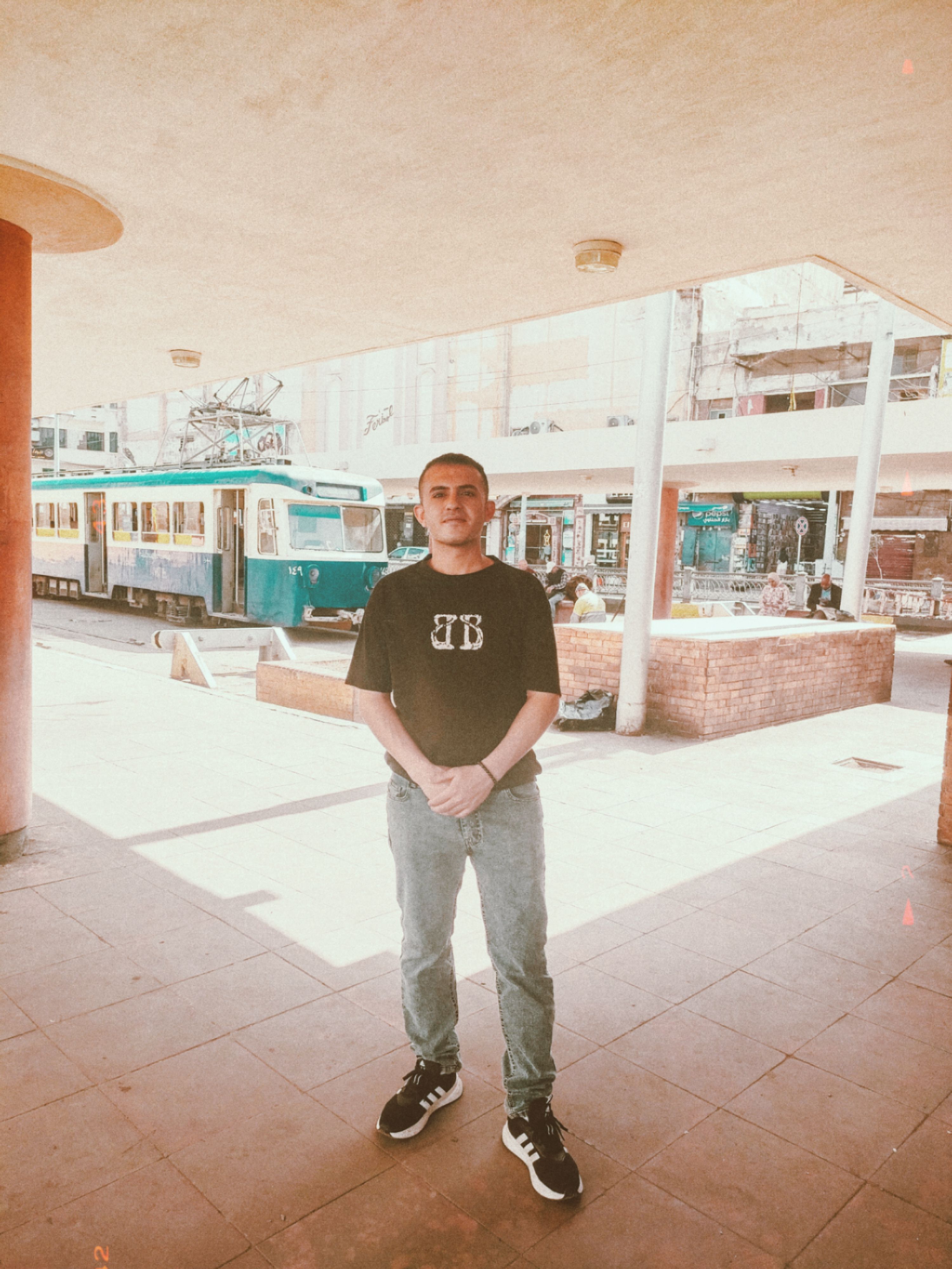 A man stands in sunlight near a blue tram, captured in a vintage filter for an urban aesthetic.