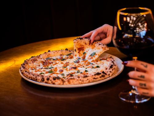 Free A hand reaches for a slice of gourmet cheese pizza paired with red wine, creating an upscale dining ambiance. Stock Photo
