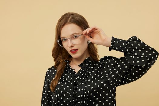 A fashionable woman wearing eyeglasses and a polka dot blouse poses against a beige background.