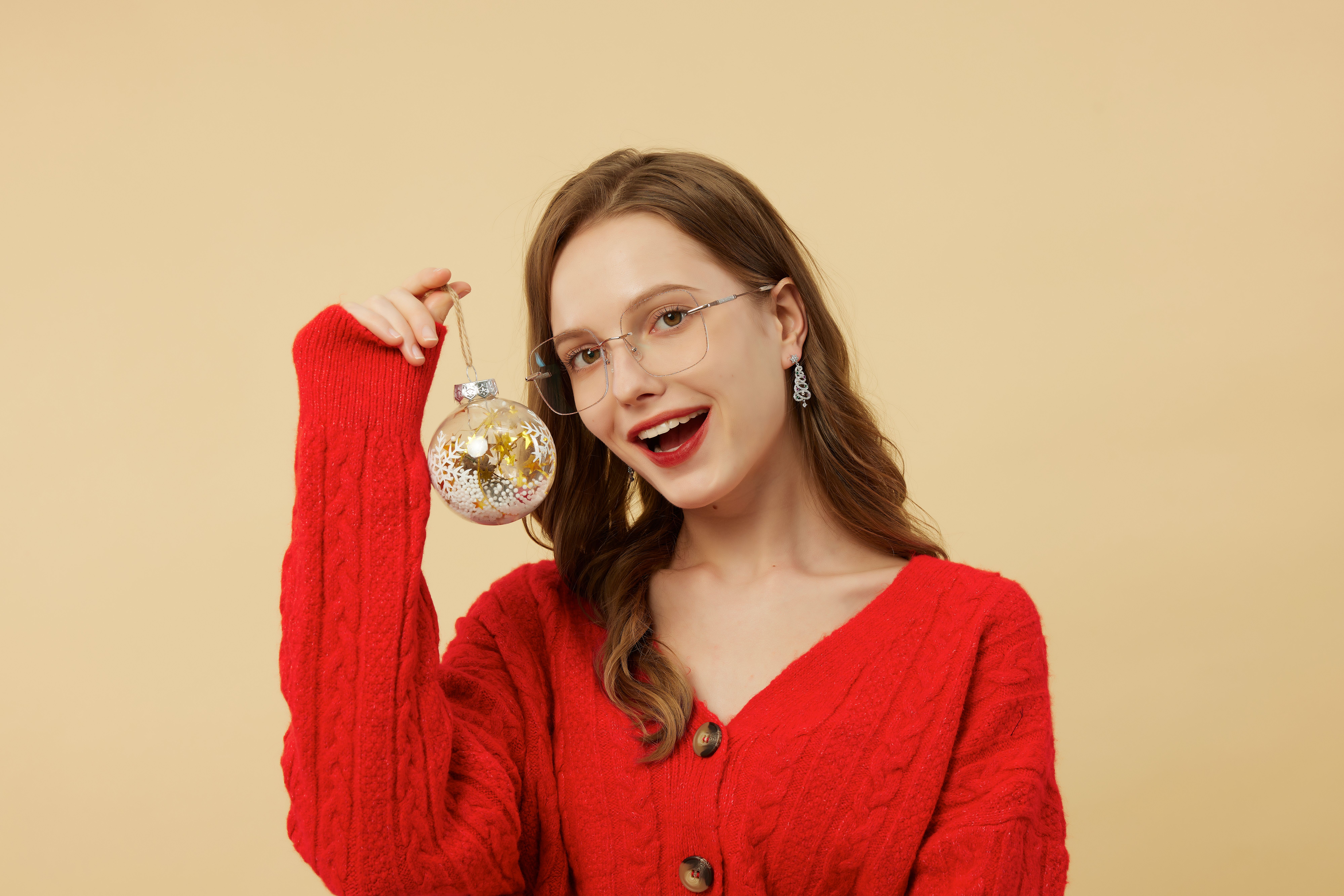 Femme souriante dans un pull rouge tenant un ornement festif sur un fond neutre.