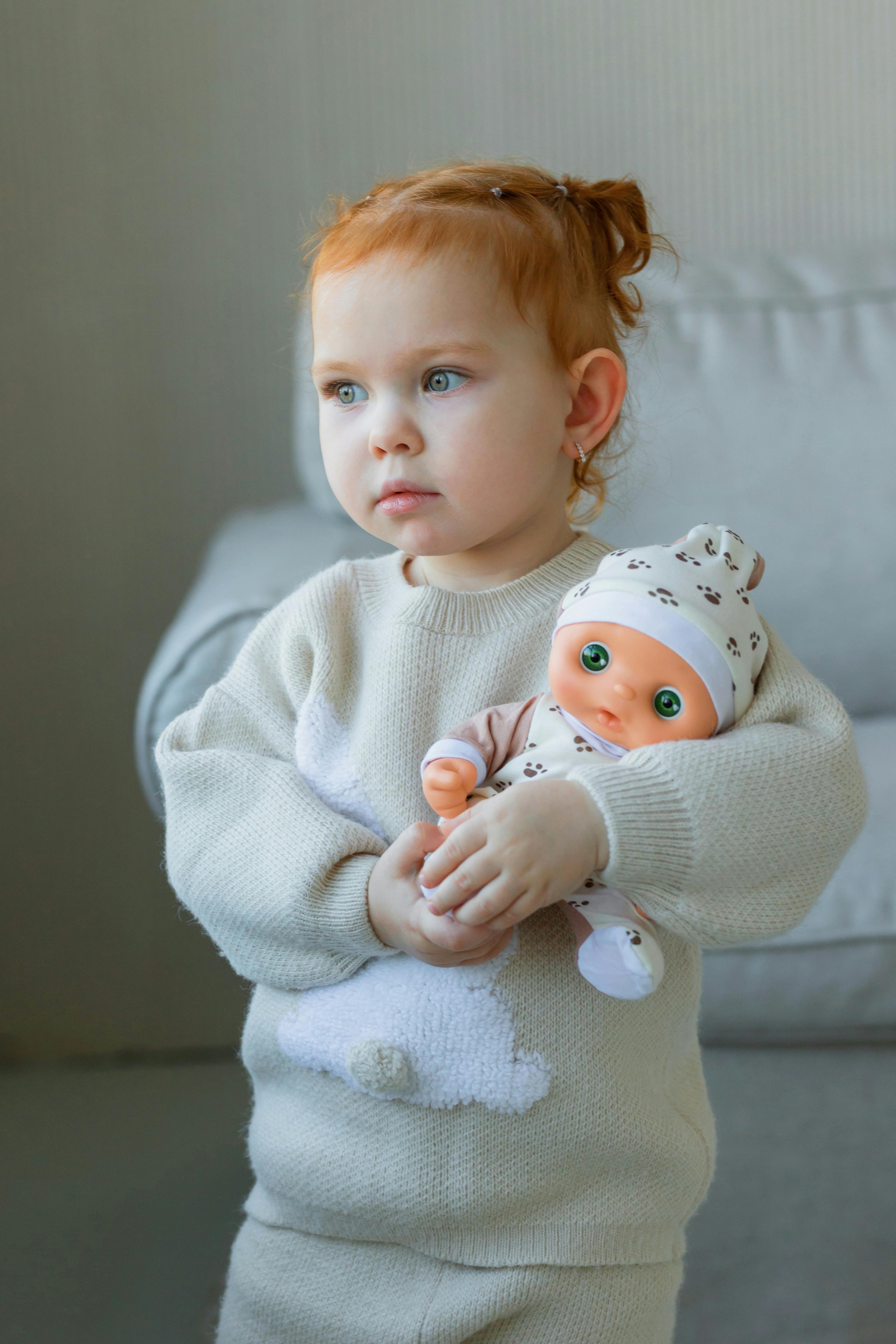 A young child with red hair tenderly holds a baby doll in a warm indoor setting.