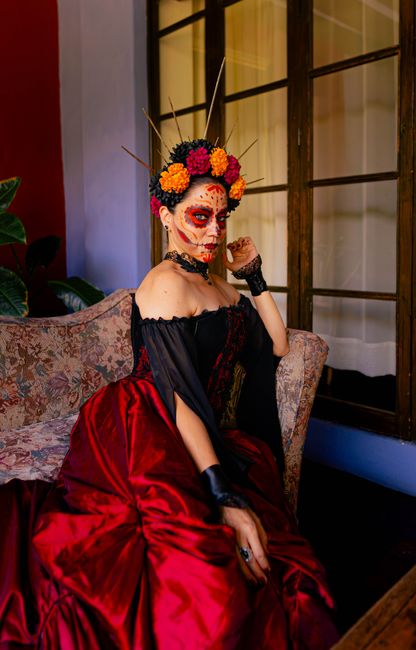 A woman in vibrant Day of the Dead makeup and attire sitting elegantly indoors.