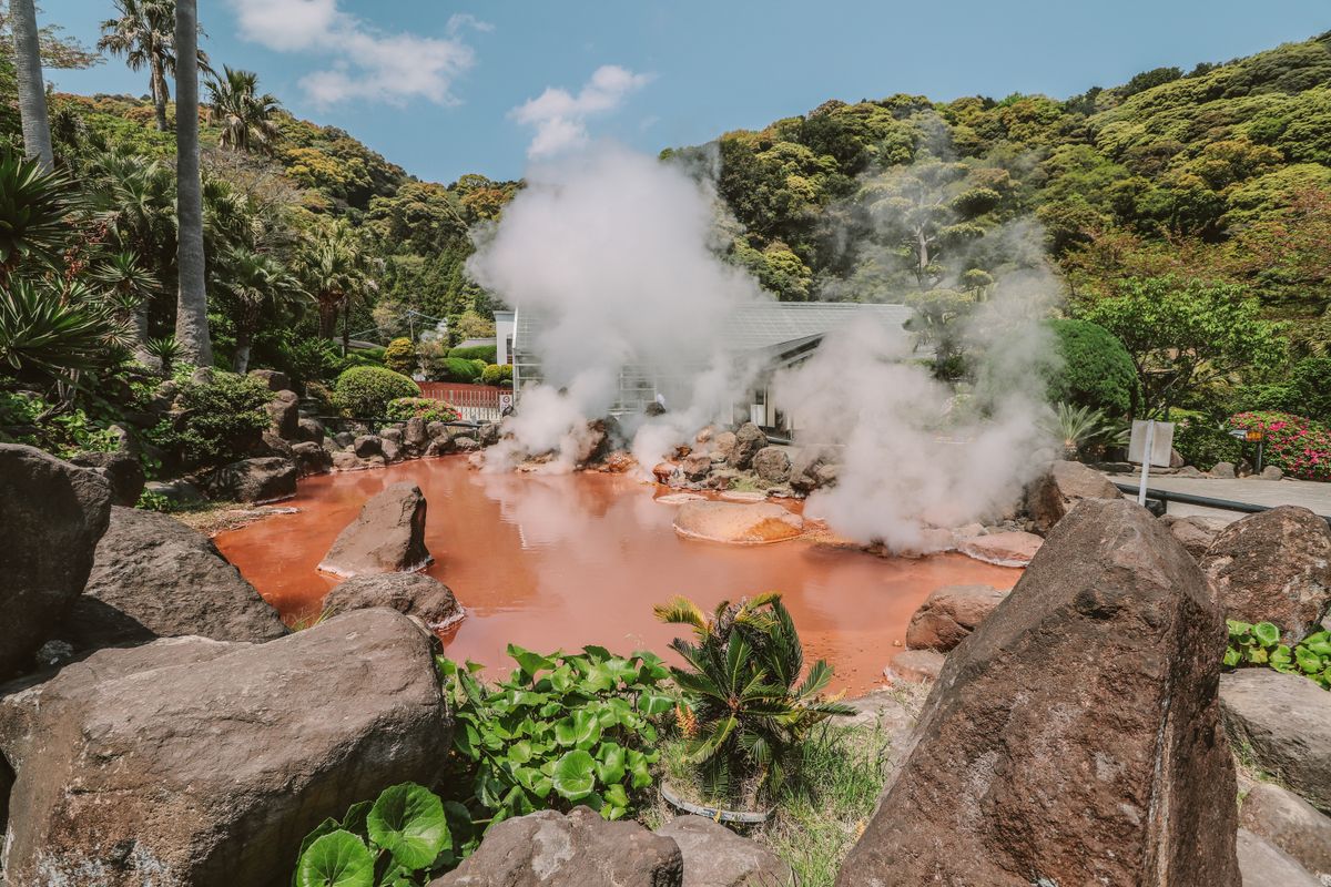 Chinoike Jigoku (Blood Pond Hell) at Beppu — the blood-red boiling pool surrounded by lush greenery with steam rising against blue sky