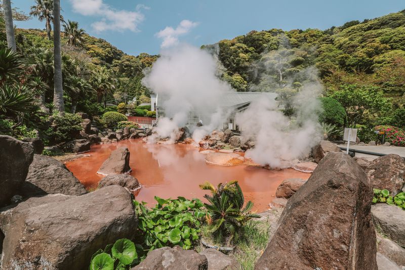 Chinoike Jigoku (Blood Pond Hell) at Beppu — the blood-red boiling pool surrounded by tropical vegetation, steam rising under open sky