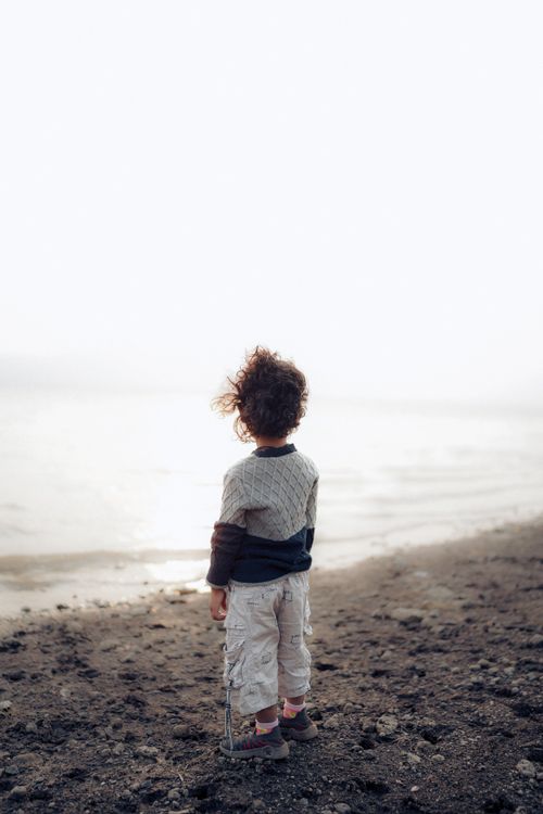 Free Child stands with curly hair, gazing at calm sea in serene setting, evoking tranquility. Stock Photo
