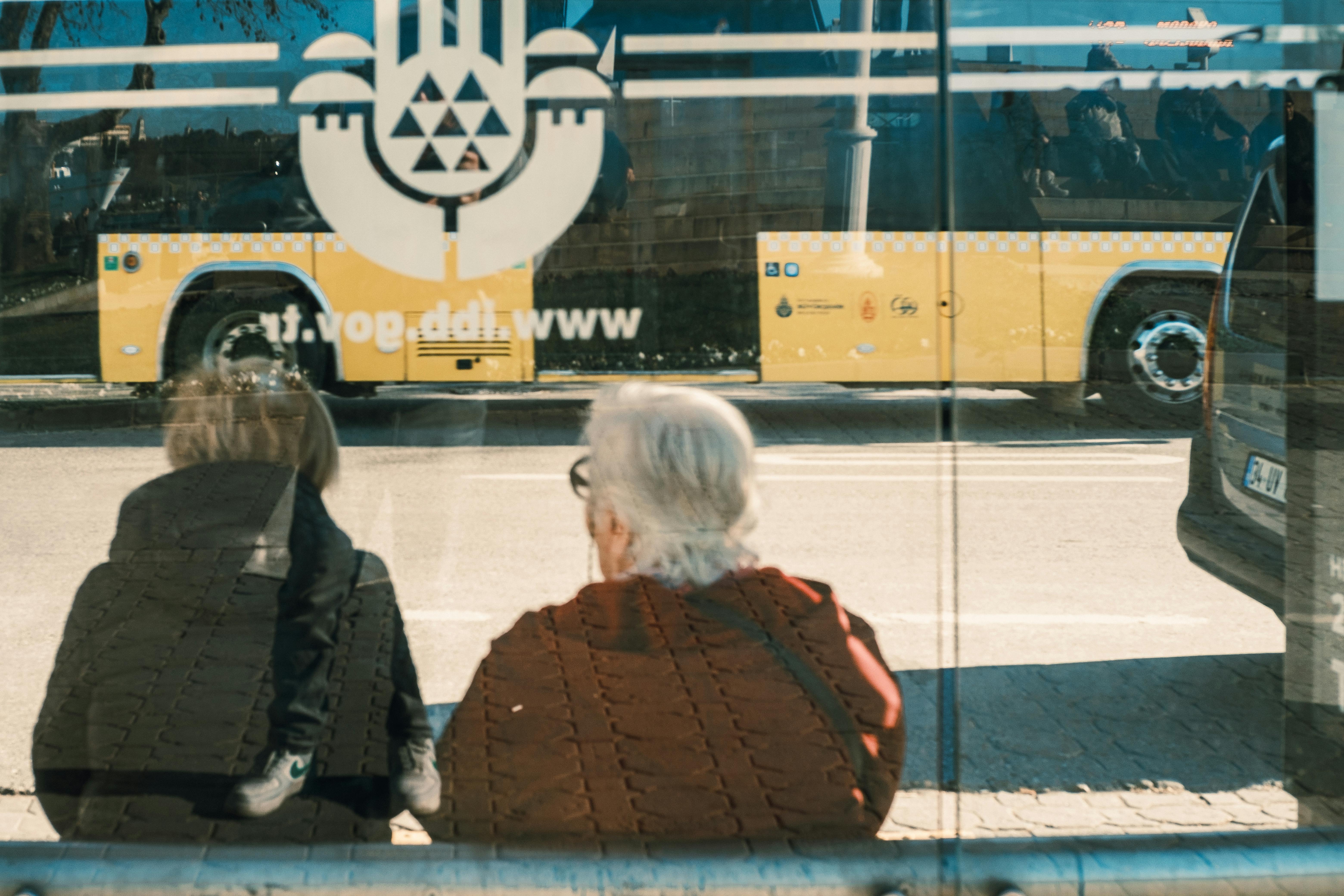 Two individuals at a bus stop with a yellow bus reflection creating an urban scene.