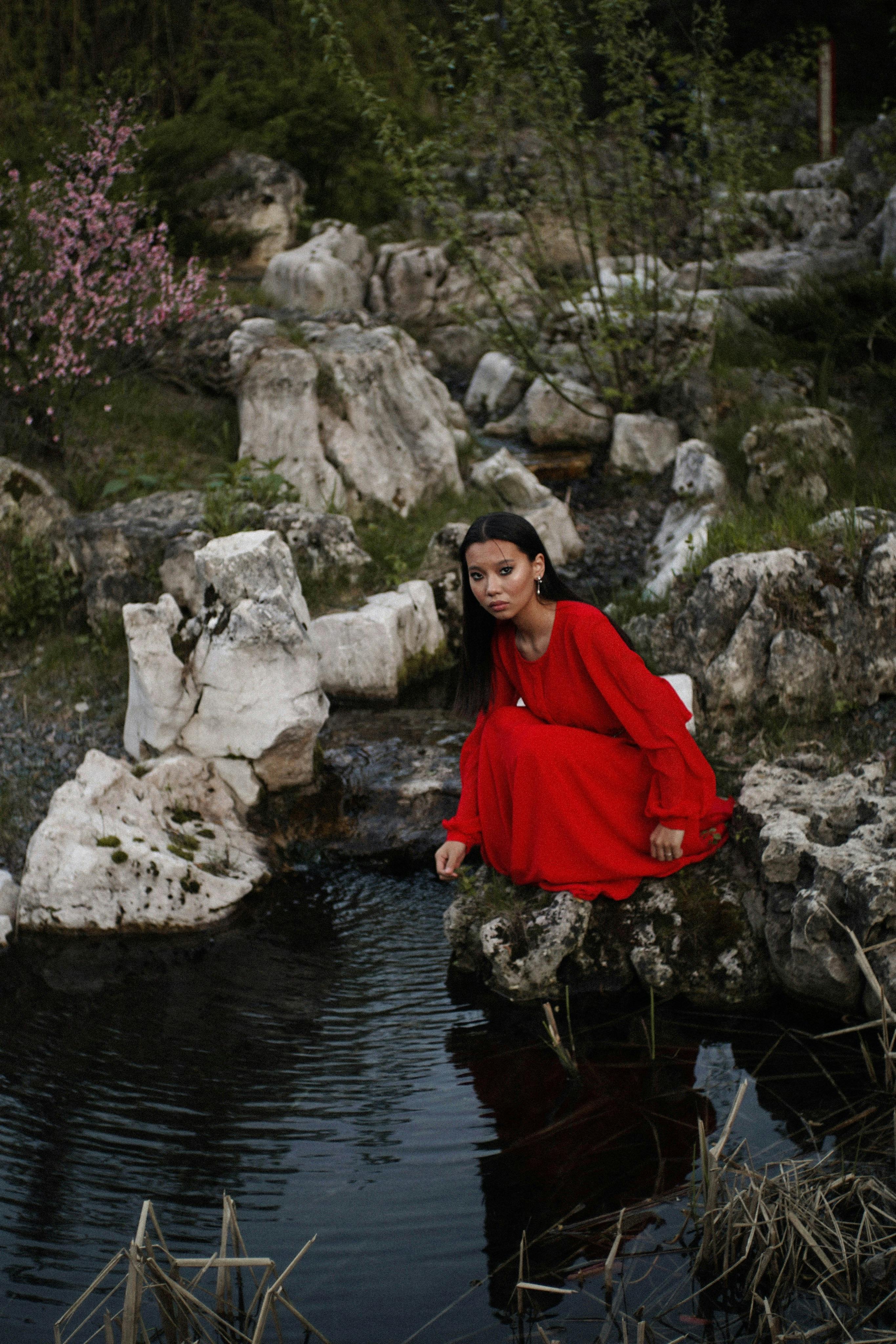 Free Woman in a flowing red dress sitting by a serene rocky pond in nature. Stock Photo