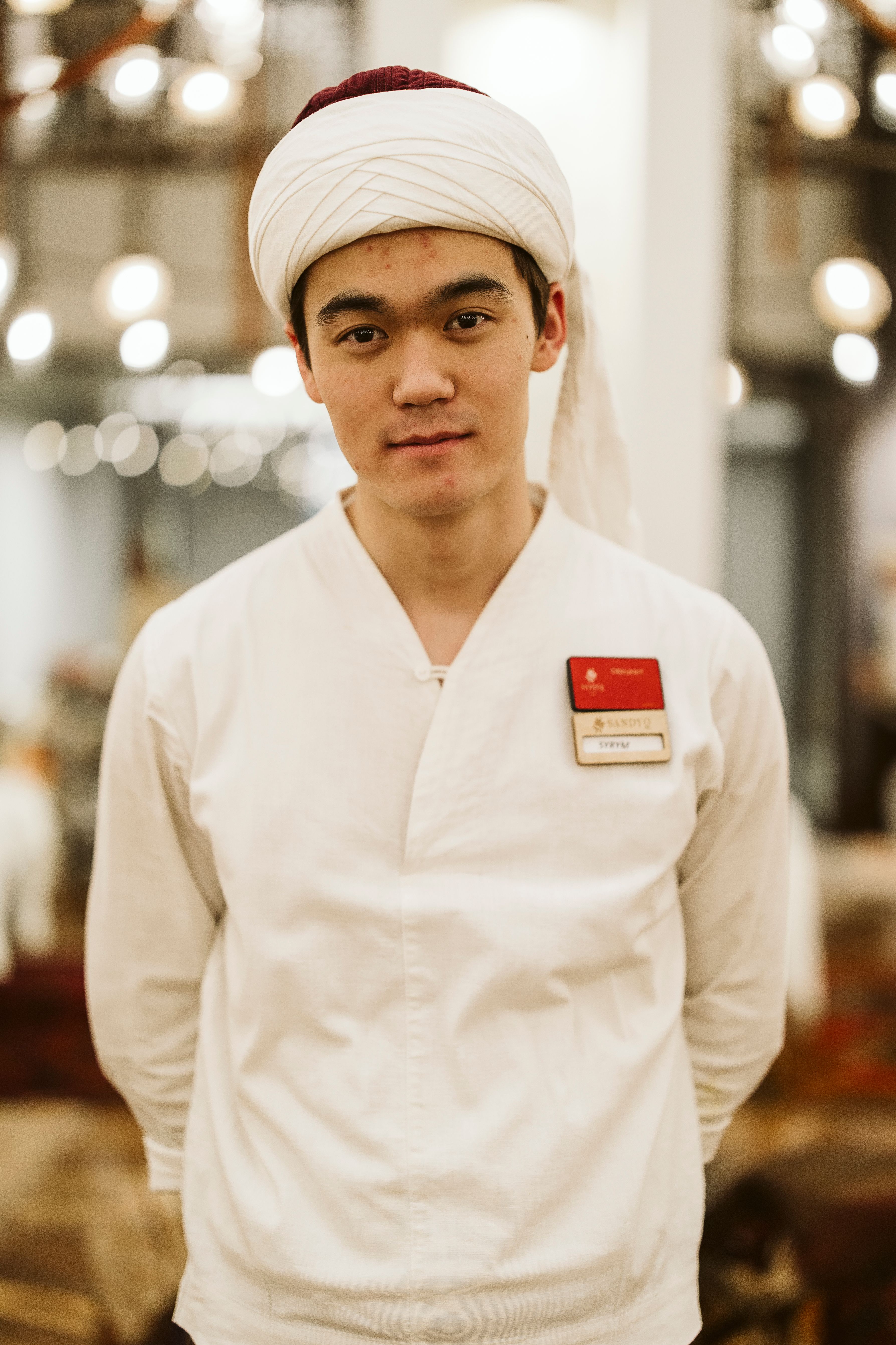 A young man in traditional attire poses indoors with a name badge, creating a cultural ambiance.