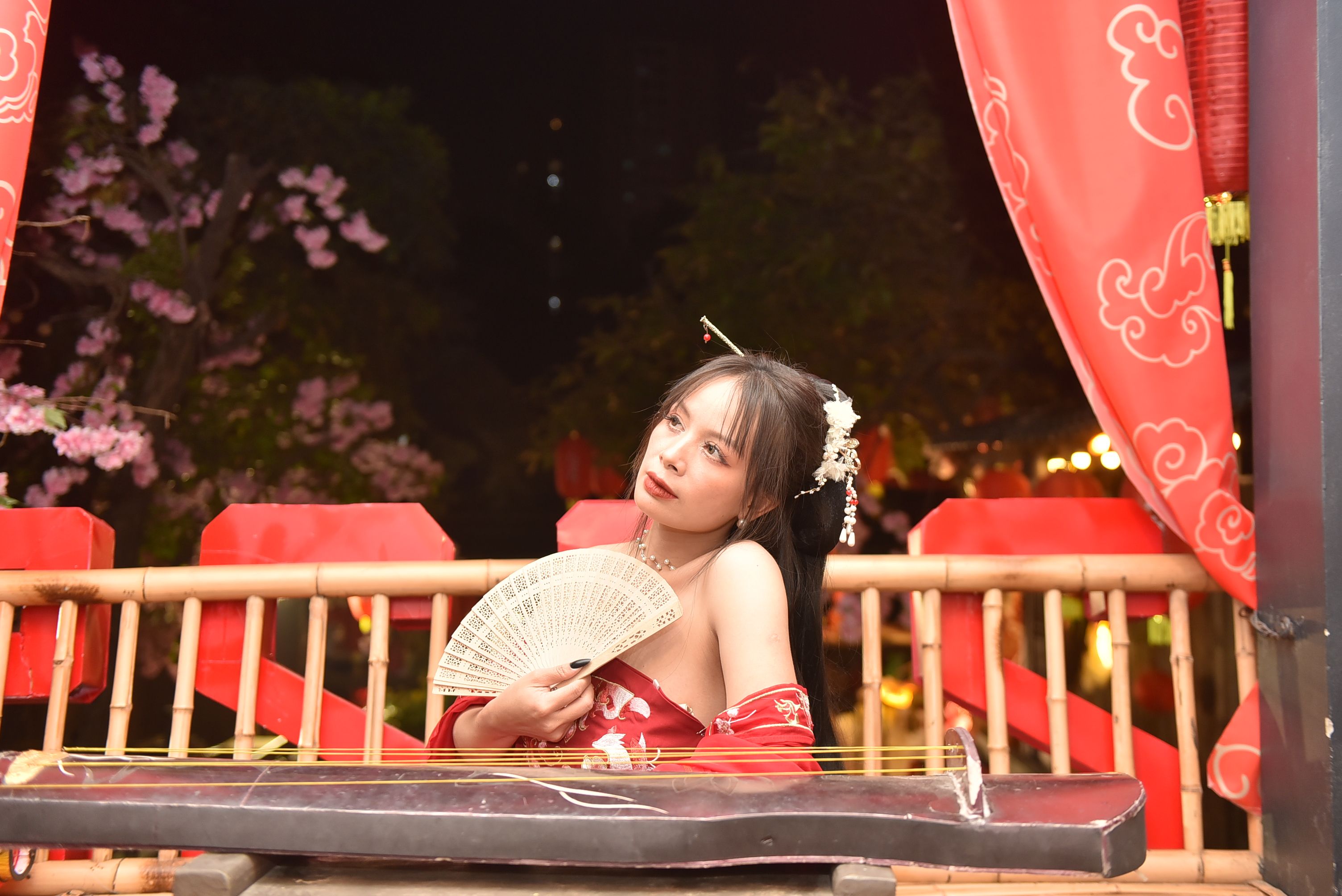 A woman in traditional attire plays the guzheng at night with cherry blossoms.