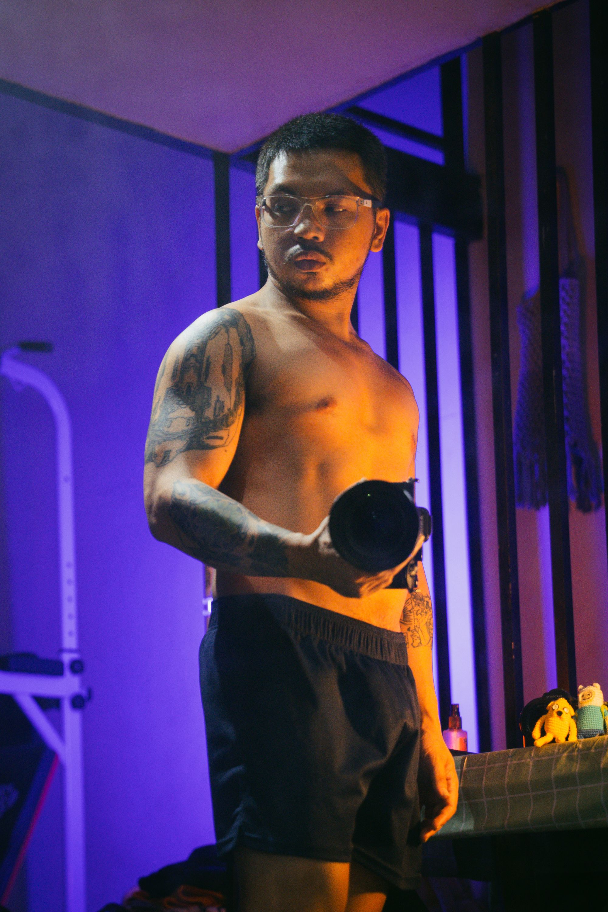 A tattooed man holding a flashlight stands in a dimly lit room, creating a moody atmosphere.