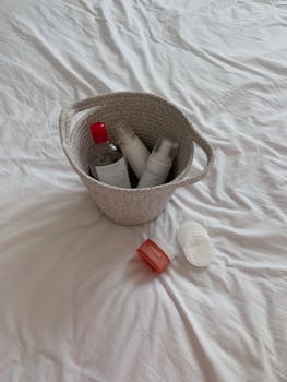 A collection of skincare products elegantly arranged in a woven basket on white bed sheets.