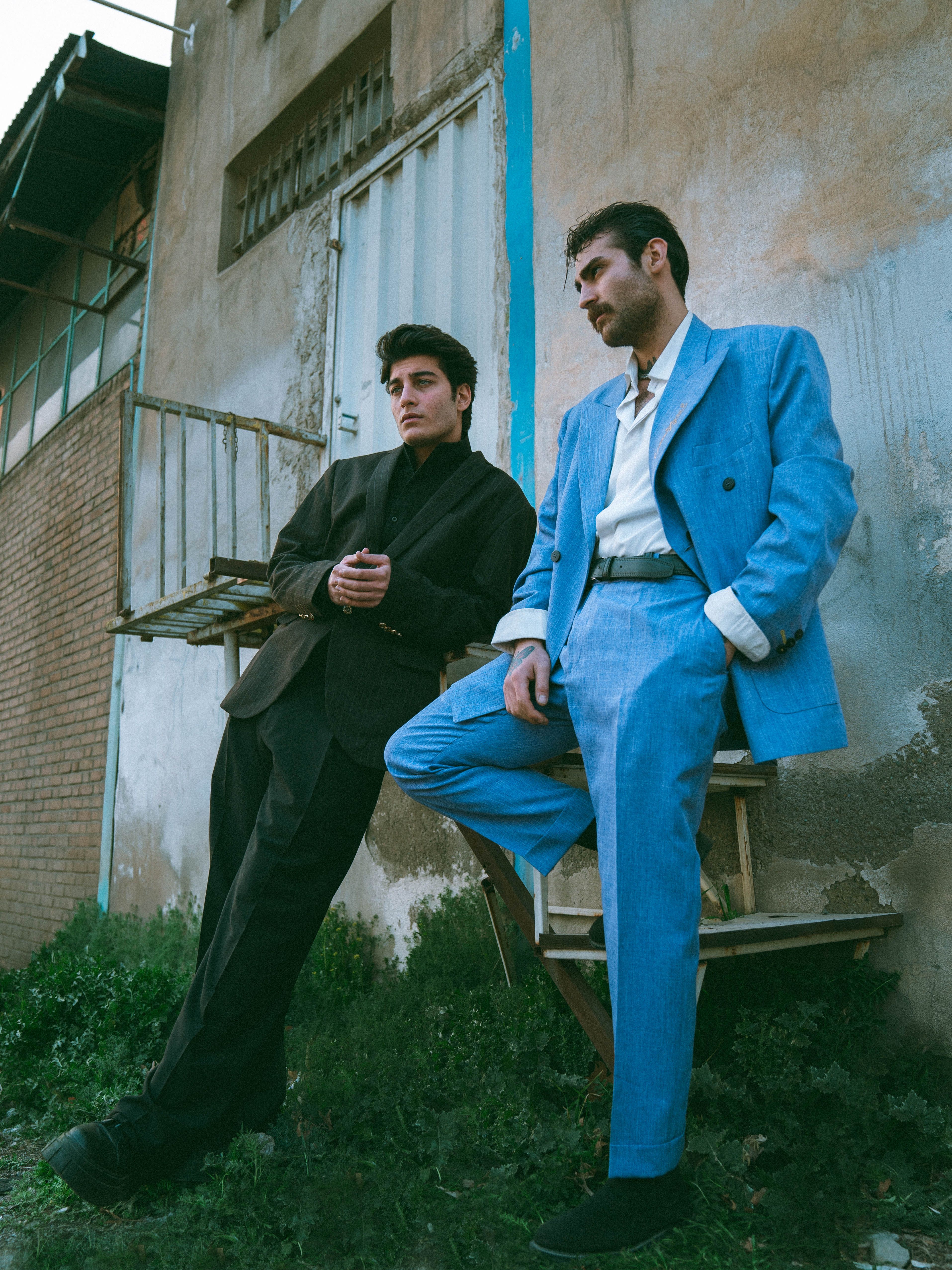 Two men in vintage suits pose dramatically outdoors against a rustic building.