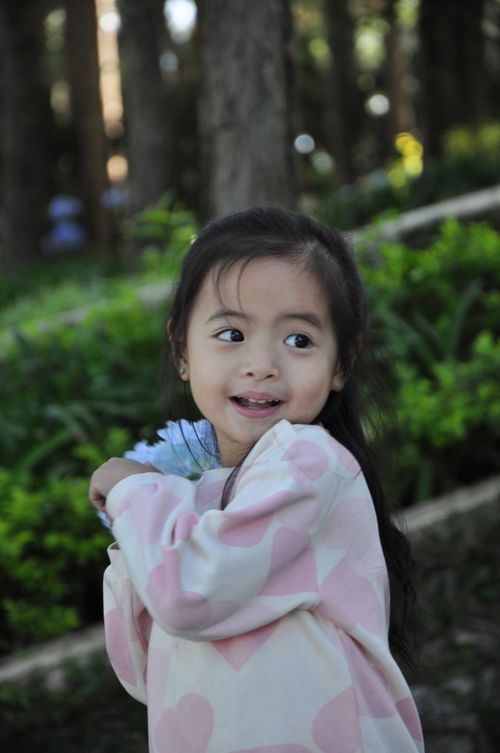 Free Cute Asian baby girl smiling in an outdoor portrait, surrounded by lush greenery. Stock Photo