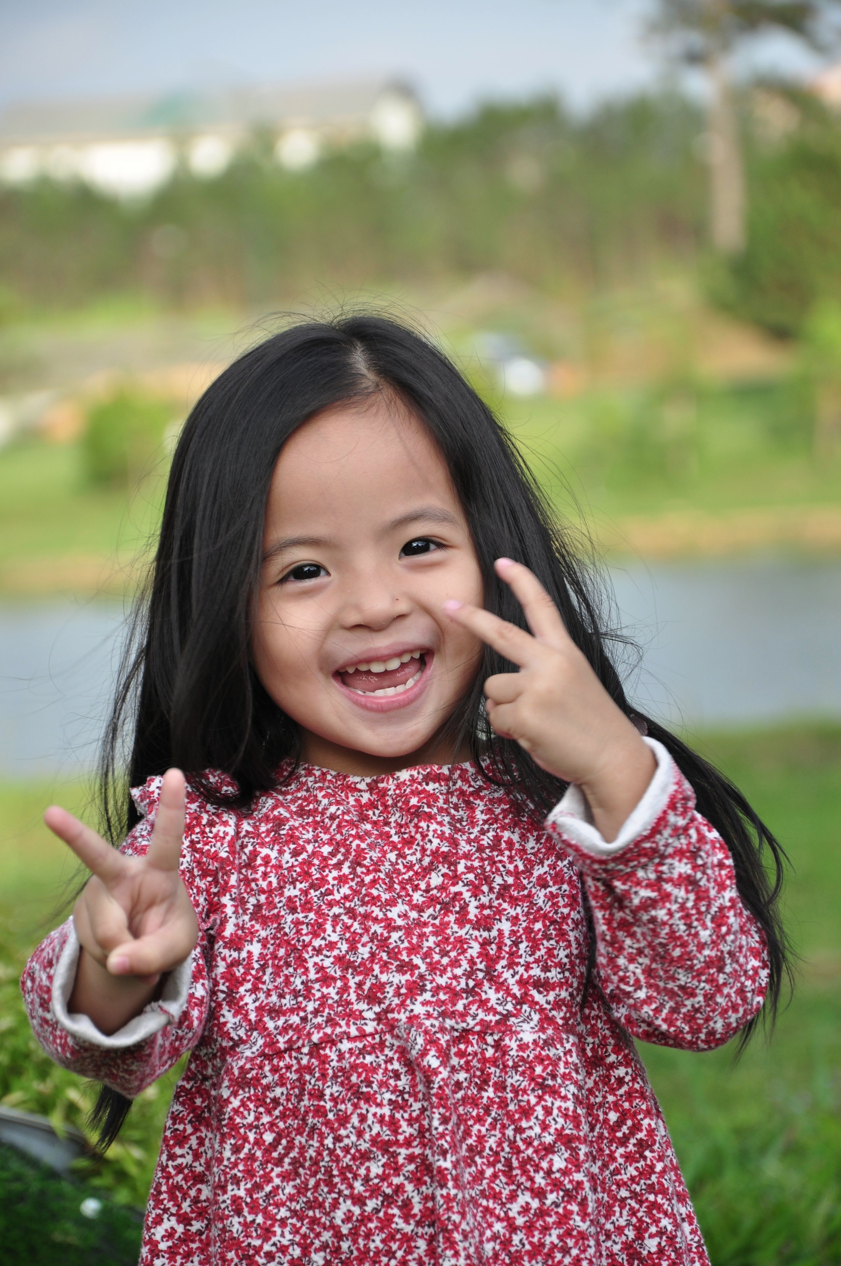 Smiling Asian girl making peace signs in a red floral dress outdoors by a lake. Joyful and playful child portrait.